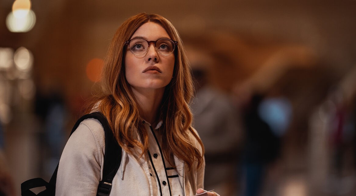 Das Titelbild zu den besten Filmen und Serien mit Sydney Sweeney zeigt die junge Schauspielerin in dem Film Madame Web. Sie steht mit einem Rucksack über der Schulter in einem Raum und blickt nach oben. Der Hintergrund ist vollkommen verschwommen und man erkennt nur Lichter am linken Bildrand. Sweeney trägt eine Brille und ihre langen, blonden Haare fallen über ihre Schultern. Sie trägt eine hellgraue Sweatshirtjacke und darunter ein schwarz-weißes Hemd. Am unteren Bildrand ist zu erkennen, dass sie etwas in ihren Händen hält, ein kleines Kärtchen. Ihr Blick nach oben sieht konzentiert aus, als würde sie eine Anzeigetafel lesen oder ähnliches.