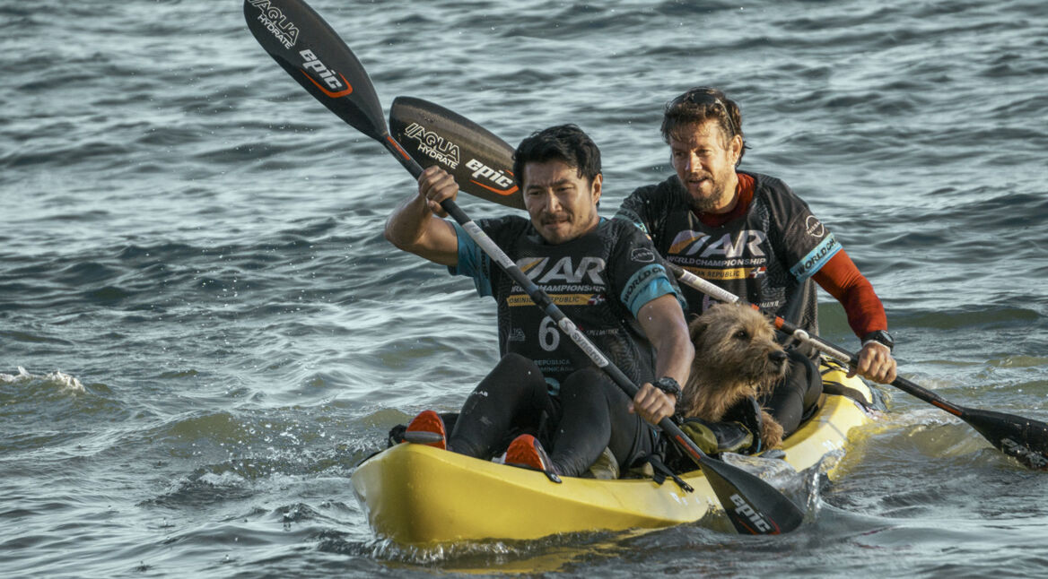 Auf diesem Bild zu den GigaTV Highlights im August sind die Schauspieler Mark Wahlberg (hinten) und Simu Liu (vorn) in einer actionreichen Szene im Film "Arthur der große" zu sehen. Sie paddeln gemeinsam in einem gelben Kajak über unruhiges Wasser, während ein kleiner Hund vorne im Kajak sitzt. Beide tragen Sportkleidung und wirken konzentriert, während sie die Wellen meistern. Die Szene vermittelt Abenteuer und Teamarbeit, perfekt passend zu den aufregenden Inhalten der GigaTV Highlights im August.