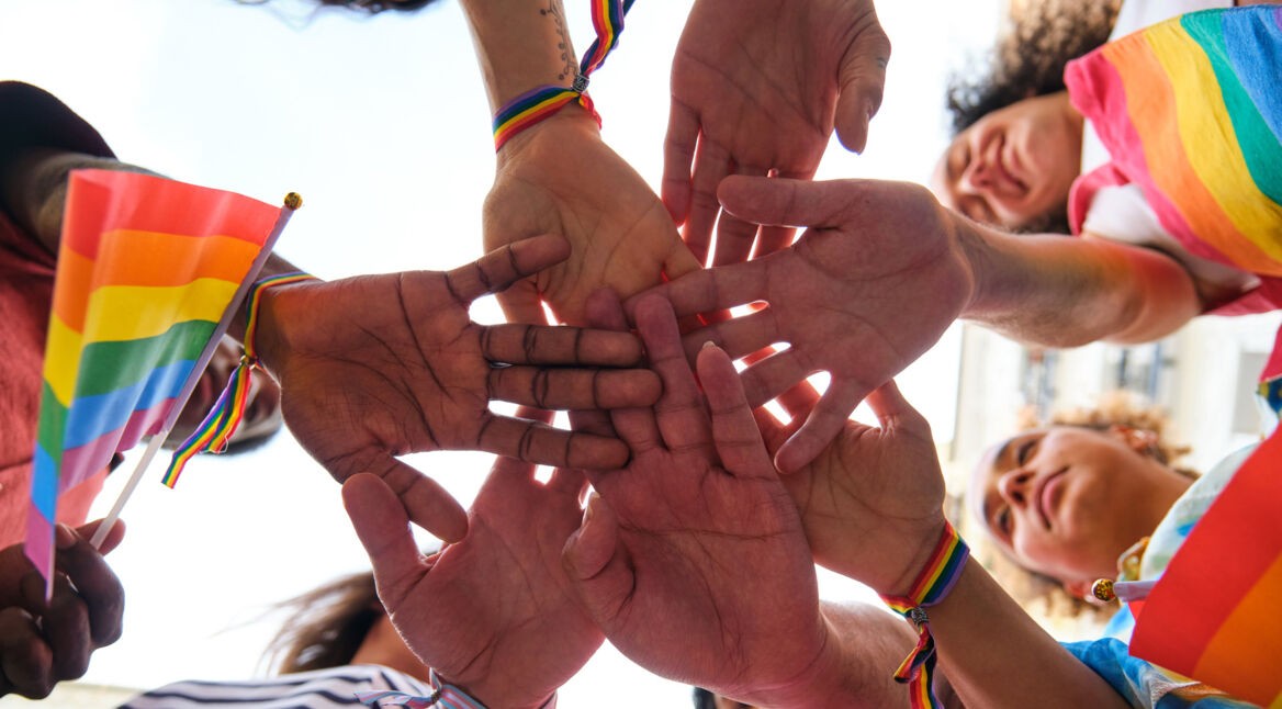 Das Bild zum International Ally Day zeigt eine Gruppe von Menschen, die ihre Hände in der Mitte zusammenschlagen. Sie tragen Armbänder und Kleidung in Regenbogenfarben und halten eine Regenbogenflagge. Die Gesichter der Personen sind fröhlich und motiviert. Das Bild symbolisiert Gemeinschaft und Unterstützung. Keyword: International Ally Day.