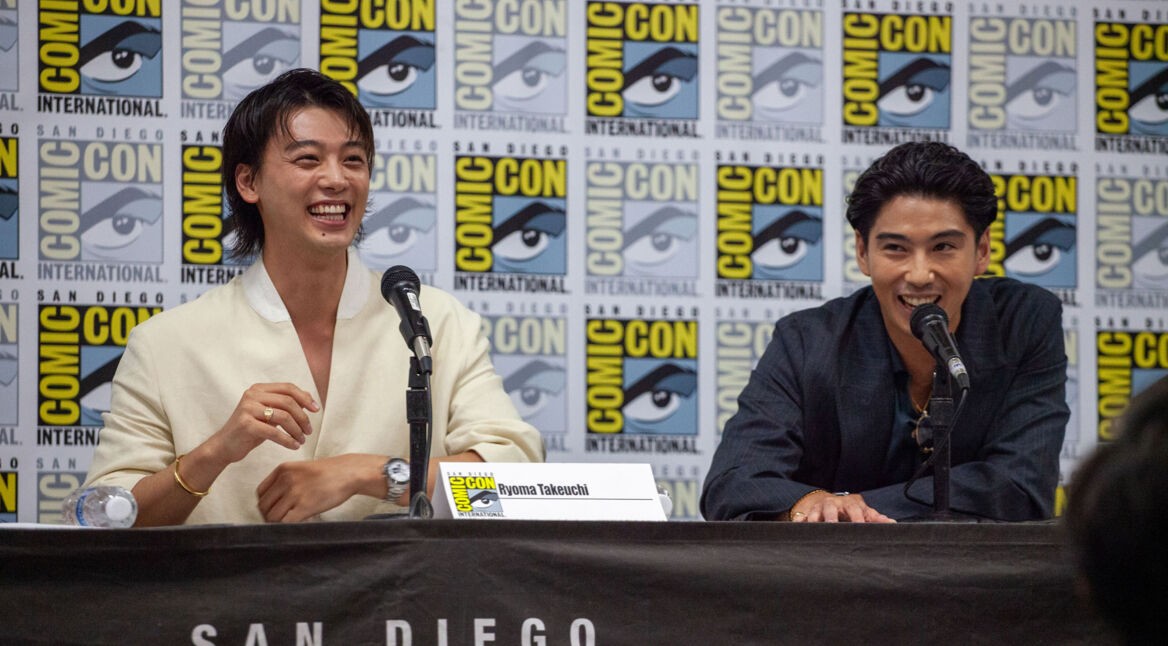 Auf diesem Bild von der Comic-Con sind die Schauspieler Ryoma Takeuchi (links) und Kento Kaku (rechts) bei einem Panel zu sehen. Beide lachen und wirken entspannt, während sie vor einem Hintergrund mit Comic-Con-Logos sitzen. Takeuchi trägt ein weißes Hemd, während Kaku in einem dunklen Anzug gekleidet ist. Sie sprechen über ihre Rollen in der "Like a Dragon: Yakuza"-Serie.