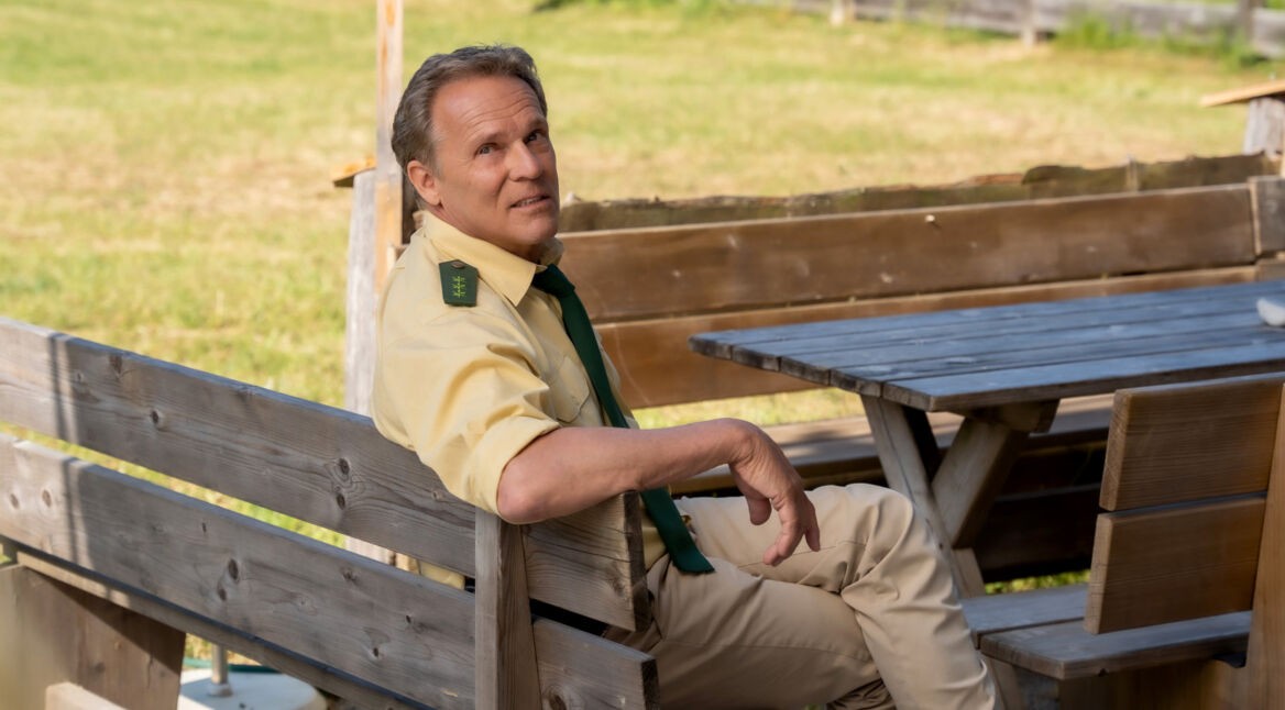 Polizeiobermeister Franz Hubert aus der Serie "Hubert und Staller" sitzt auf einer Bank im Grünen.