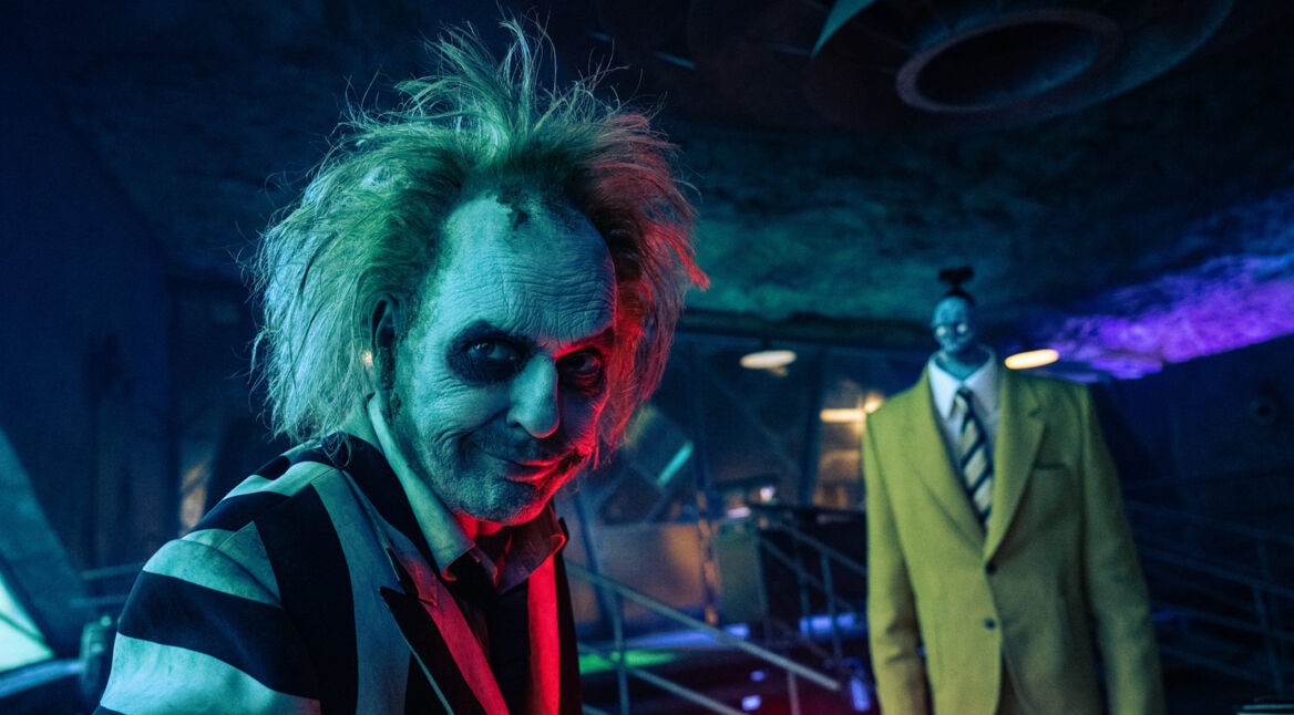 Auf dem Bild zu "Beetlejuice Beetlejuice Ende erklärt" ist Michael Keaton als der exzentrische Poltergeist Beetlejuice in seinem ikonischen schwarz-weiß gestreiften Anzug zu sehen. Sein zerzaustes Haar und das düstere Make-up verstärken den skurrilen, geisterhaften Look. Im Hintergrund steht eine düstere, große Figur in einem gelben Anzug, die düstere Stimmung der Szene wird durch das kalte, bläuliche Licht unterstrichen. Die Szene wirkt mysteriös und unheimlich.