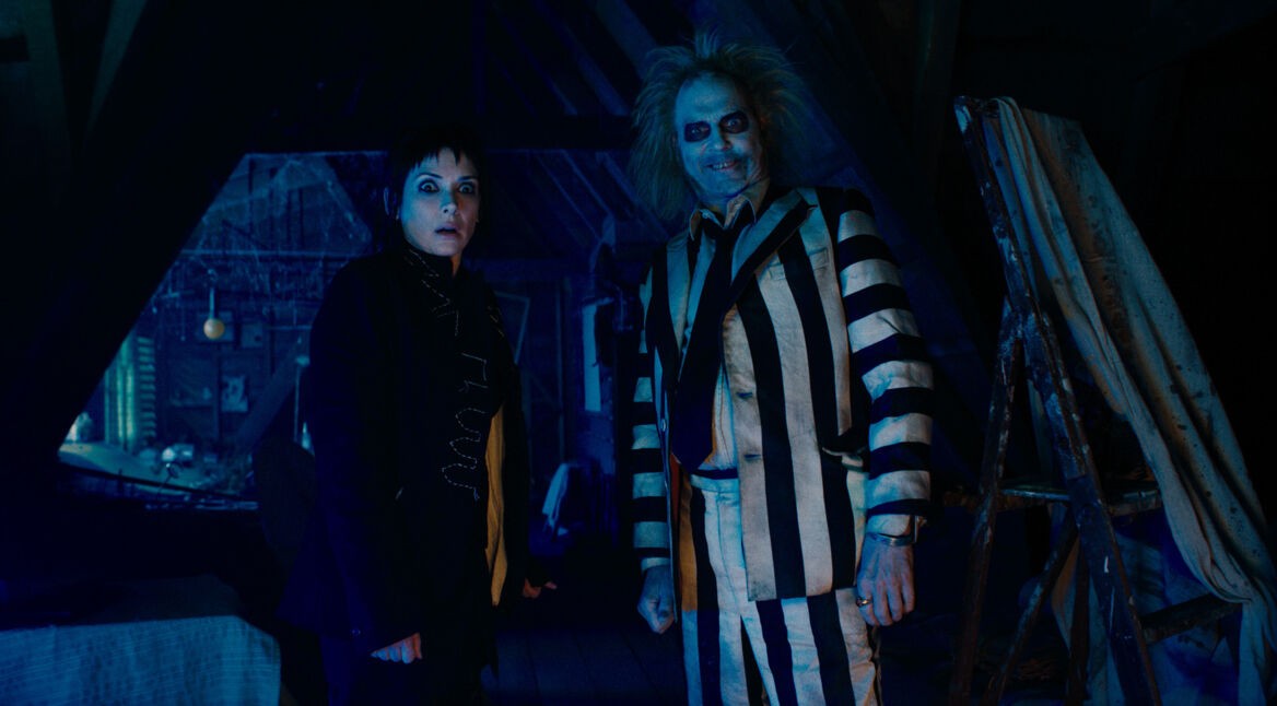 Auf diesem Bild zur Beetlejuice Beetlejuice Kritik stehen Winona Ryder als Lydia und Michael Keaton als Beetlejuice nebeneinander in einem düsteren, von Spinnweben bedeckten Dachboden. Lydia trägt schwarze Kleidung und blickt überrascht in die Kamera, während Beetlejuice in seinem markanten schwarz-weiß gestreiften Anzug grinst. Die Szene ist in kaltes, blaues Licht getaucht, was eine geheimnisvolle und leicht unheimliche Atmosphäre erzeugt. Im Hintergrund sind ein Leitergerüst und Abdeckungen zu erkennen.