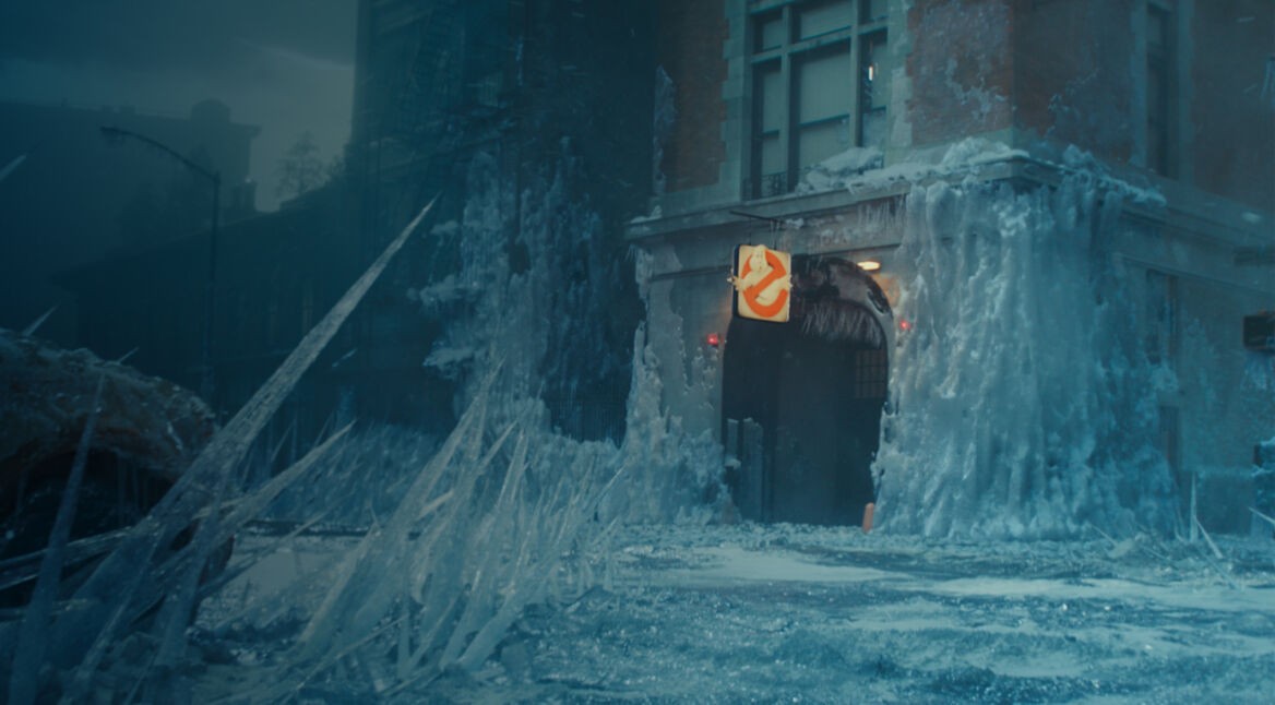 Auf dem Bild aus dem Film Ghostbusters Frozen Empire ist das legendäre Ghostbusters-Hauptquartier in einer verschneiten, eingefrorenen Stadt zu sehen. Über dem Eingang hängt das ikonische Ghostbusters-Logo, während Eiszapfen und gefrorene Strukturen die Umgebung bedecken. Die düstere Atmosphäre deutet auf eine Bedrohung hin. Es ist wahrscheinlich, dass das Gebäude auch bei der Ghostbusters-Serie von Netflix eine Rolle spielt.