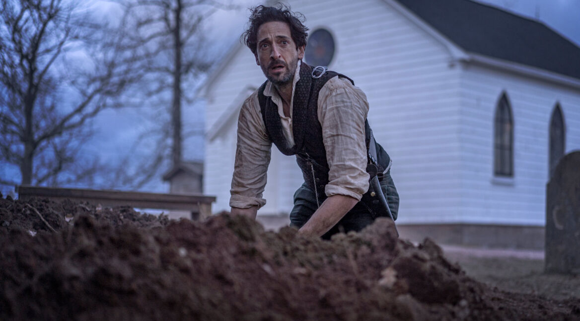 Auf dem Bild zu den GigaTV Highlights im Oktober ist Adrien Brody als Charles Boone in einer dramatischen Szene aus "Chapelwaite" zu sehen. Er kniet vor einem frisch ausgehobenen Grab, umgeben von dunkler Erde, mit einem besorgten Blick. Im Hintergrund steht eine weiße Kirche vor einem düsteren Himmel, was die unheilvolle Stimmung unterstreicht. Seine Kleidung ist zerknittert und wirkt durch die Szene strapaziert.