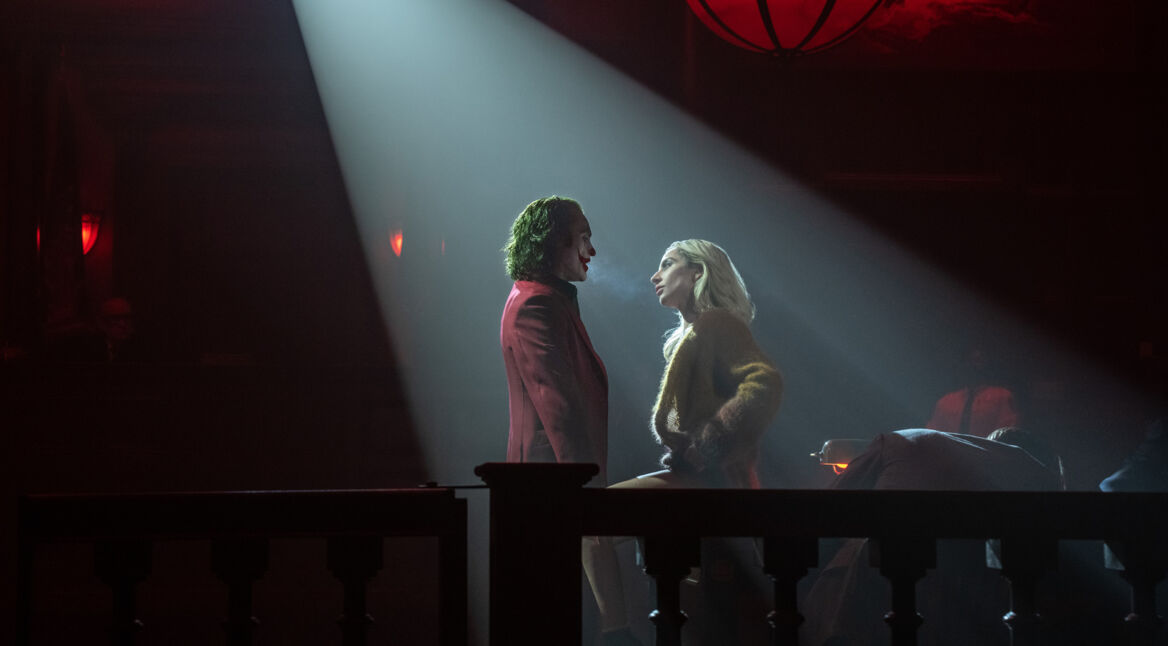 Auf dem Bild zum Faktenartikel über DCs Joker sind Joaquin Phoenix als der Joker und Lady Gaga als Harley Quinn in einer intensiven Szene zu sehen. Der Joker trägt einen roten Anzug, während Harley Quinn in einem pelzähnlichen Mantel gekleidet ist. Sie stehen sich nahe, während ein dramatischer Lichtstrahl auf sie fällt, was eine geheimnisvolle und spannungsgeladene Atmosphäre schafft. Beide Charaktere wirken konzentriert und ihre Körperhaltung deutet auf eine komplexe Dynamik hin.