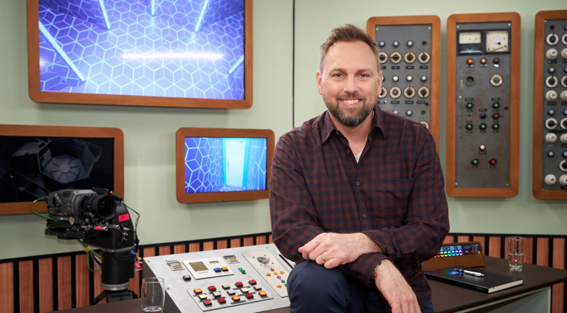 Auf dem Bild zur Prime-Video-Show "Licht Aus" ist der Moderator Steven Gätjen zu sehen, wie er lächelnd vor einer technischen Kontrolltafel sitzt. Im Hintergrund hängen mehrere Bildschirme mit blauen, geometrischen Mustern, die einen futuristischen Look verleihen. Steven trägt ein kariertes Hemd und wirkt entspannt und professionell. Die Szene vermittelt eine moderne, technikbasierte Atmosphäre.