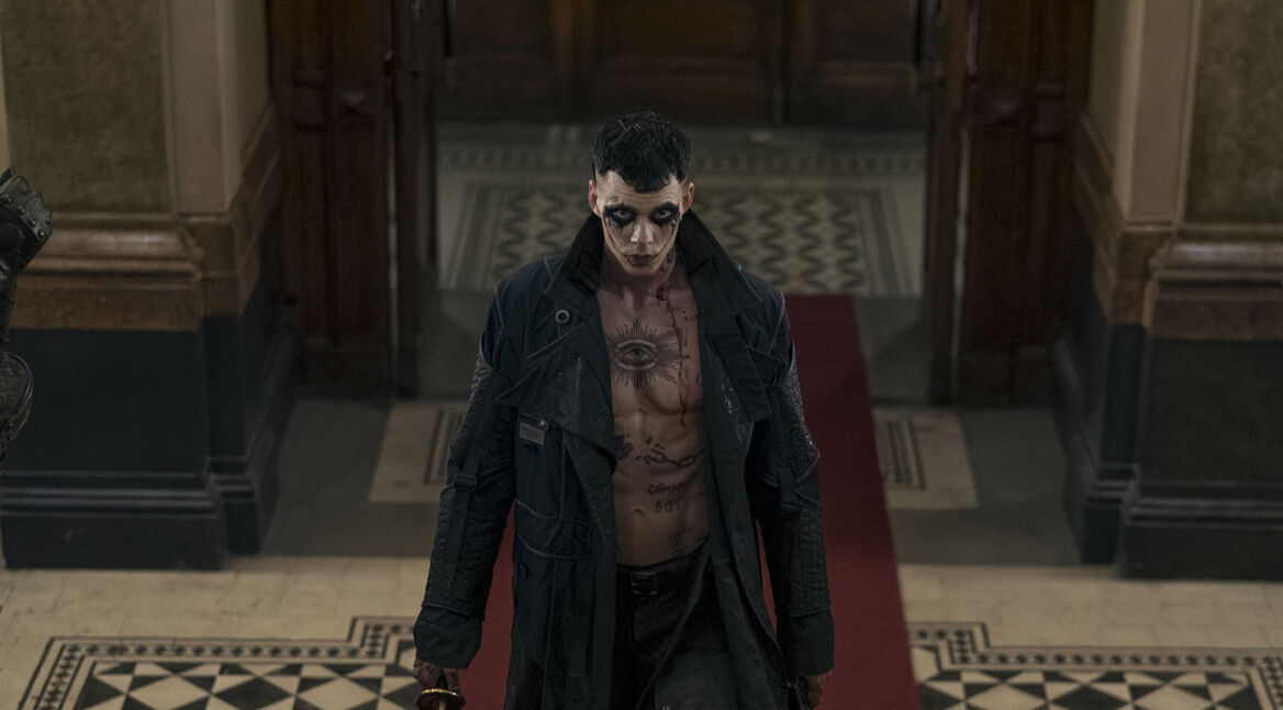 Auf dem Artikelbild zur The Crow-Kritik ist Bill Skarsgård in seiner Rolle als The Crow zu sehen. Er steht in einem alten Gebäude auf einem roten Teppich und trägt einen langen, dunklen Mantel. Sein Oberkörper ist nackt und mit mehreren Tätowierungen bedeckt, darunter ein auffälliges Auge auf der Brust. Sein Gesicht ist bleich geschminkt, mit dunklen Augenringen und blutigen Spuren. Er wirkt entschlossen und bedrohlich, während er ein Schwert in der rechten Hand hält. Die düstere, unheilvolle Atmosphäre unterstreicht die düstere Stimmung des Films.