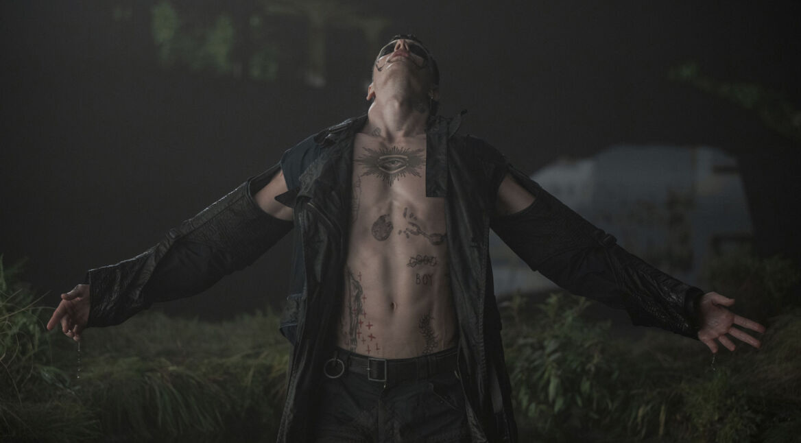 Auf dem Bild zu "The Crow Unterschiede" ist Bill Skarsgård in einer ikonischen Szene aus "The Crow (2024)" zu sehen. Mit nacktem Oberkörper und zahlreichen Tattoos, darunter ein großes Auge auf seiner Brust, steht er mit ausgebreiteten Armen in einer düsteren, regnerischen Umgebung. Sein Gesicht ist teilweise schwarz bemalt, während er den Kopf nach hinten lehnt, als würde er einen intensiven Moment durchleben. Die Szene strahlt eine düstere, fast mystische Atmosphäre aus, passend zur Ästhetik des Films.