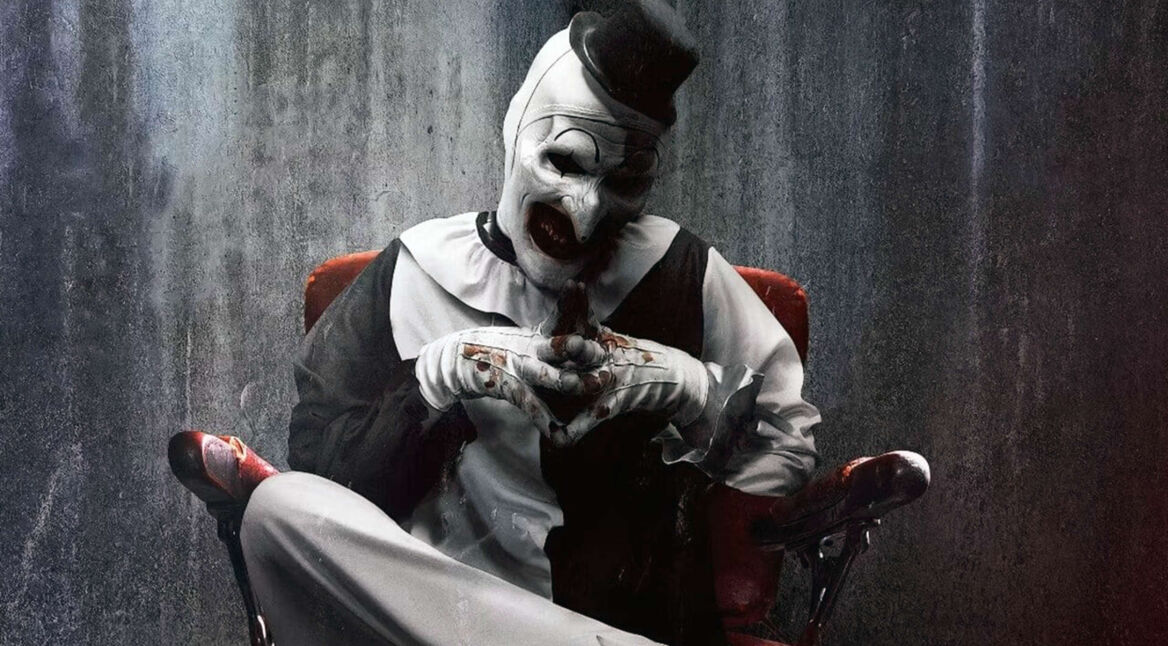 Art the Clown aus dem Film "Terrifier 3" sitzt mit überschlagenen Beinen auf einem Stuhl vor einer Betonwand.