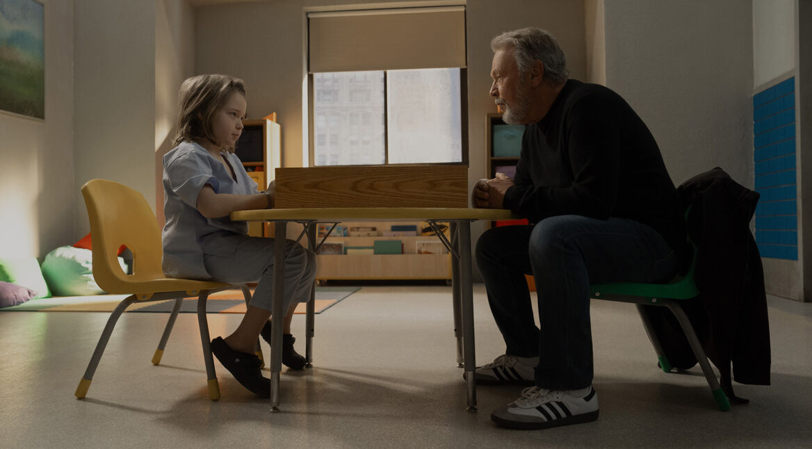 In einer Szene aus der Before-Serie auf Apple TV+ sitzen Noah auf einem gelben Stuhl links und Eli auf einem grünen Stuhl rechts an einem kleinen Tisch in einem schlicht eingerichteten Raum. Noah schaut konzentriert auf den Tisch, während Eli ihn ruhig und nachdenklich ansieht. Die beiden befinden sich in einer ruhigen Umgebung mit bunten Kissen im Hintergrund, was auf eine intensive und bedeutungsvolle Unterhaltung zwischen den beiden Charakteren hinweist.