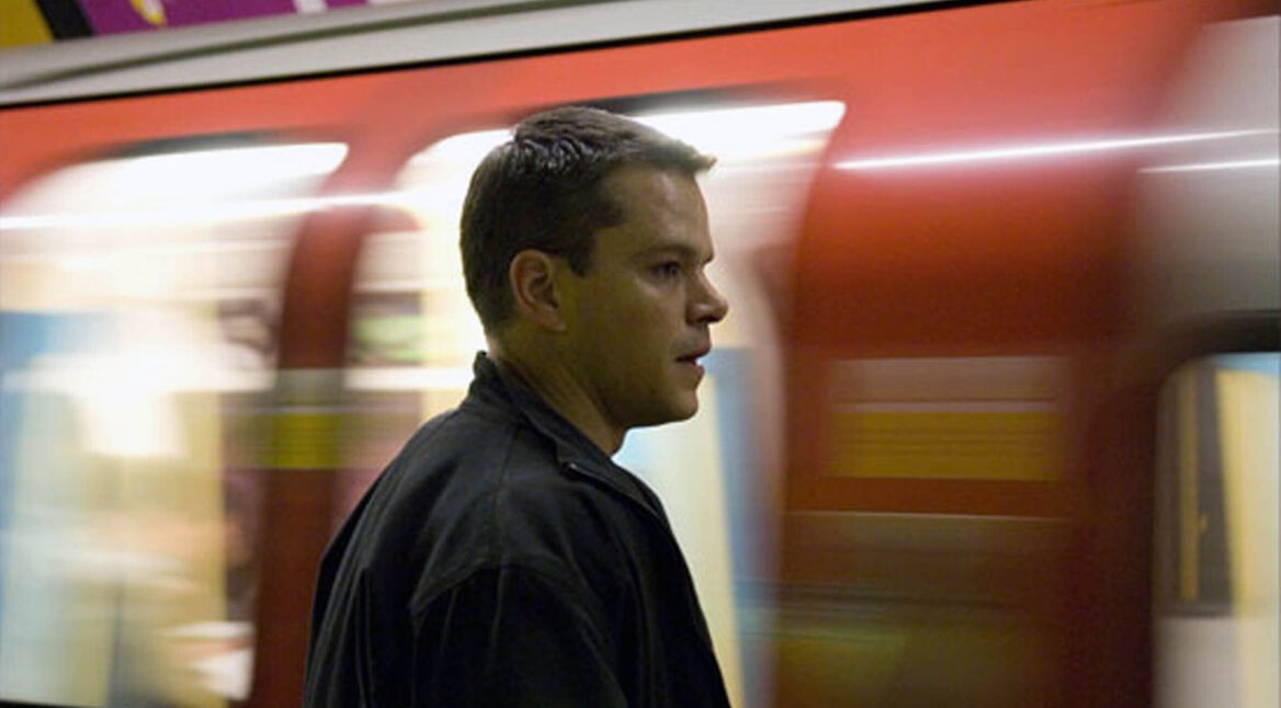 Auf dem Bild ist Jason Bourne, gespielt von Matt Damon, in einer U-Bahn-Station zu sehen. Bourne steht seitlich im Profil, fokussiert und konzentriert, während hinter ihm ein vorbeirasender roter U-Bahn-Wagen Bewegungsunschärfe erzeugt. Die Szene vermittelt Spannung und Dynamik, passend zu Bournes actionreichem Charakter und seiner ständigen Flucht.
