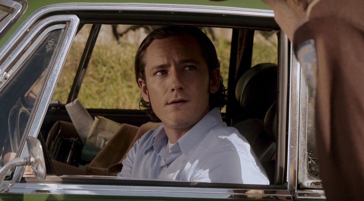 Auf dem Bild aus "Salems Lot - Brennen muss Salem" ist Lewis Pullman als Ben Mears zu sehen. Er sitzt in einem Auto, blickt nachdenklich und leicht besorgt nach oben zu einer Person, die außerhalb des Wagens steht. Seine Haltung und der ernste Ausdruck deuten auf eine angespannten Moment hin. Im Hintergrund sind grüne Bäume und Büsche zu erkennen, was die Szene ruhig wirken lässt.