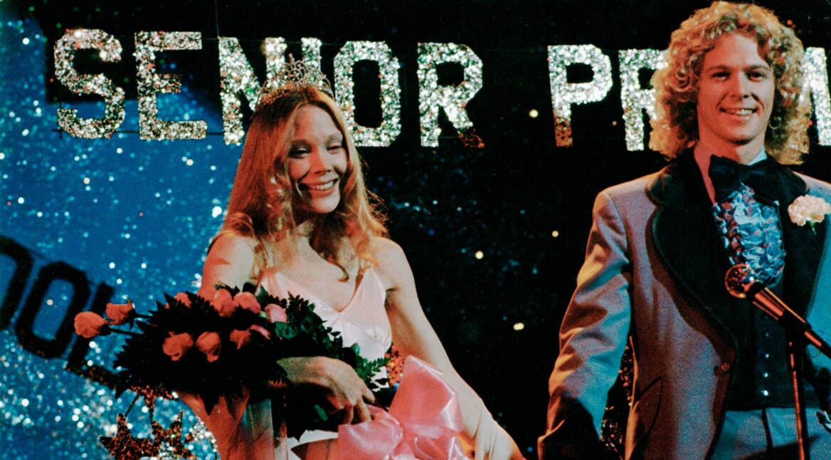 Auf dem Bild zur Serie Carrie sieht man die gekrönte Carrie, gespielt von Sissy Spacek, auf ihrem Abschlussball. Sie trägt eine Tiara und hält einen Blumenstrauß mit rosafarbenen Rosen, während sie lächelnd neben ihrem Ballkönig steht. Im Hintergrund leuchtet ein großes "Senior Prom"-Schild in funkelnden Buchstaben. Die Szene wirkt harmonisch und festlich, mit einer Spur von Spannung, die für die düstere Handlung typisch ist.