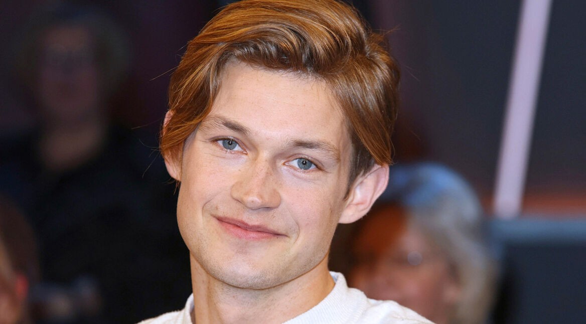 Auf dem Bild ist der deutsche Schauspieler Damian Hardung zu sehen, wie er in die Kamera lächelt. Er hat hellbraune Haare, die leicht zur Seite gestylt sind, und blaue Augen. Sein Gesichtsausdruck wirkt freundlich und entspannt. Der Hintergrund ist unscharf, sodass der Fokus auf seinem Gesicht liegt.