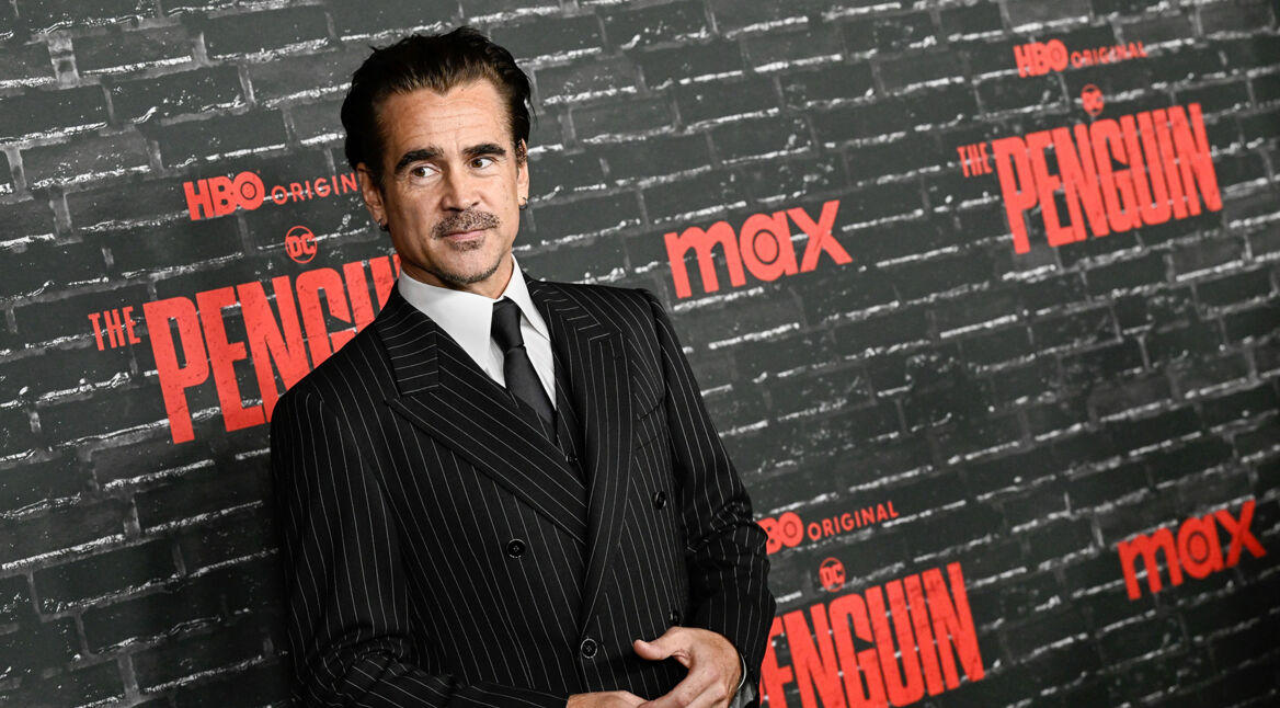 Auf dem Bild ist Colin Farrell zu sehen, der die Hauptrolle als Oswald Cobblepot, auch bekannt als der Pinguin, in der Serie "Der Pinguin" spielt. Farrell trägt einen eleganten, dunklen Nadelstreifenanzug und steht vor einer schwarzen Backsteinwand mit den roten Logos von HBO und der Serie "The Penguin". Das Bild ist auf der Premiere entstanden. Seine Haltung wirkt selbstbewusst und leicht nachdenklich.