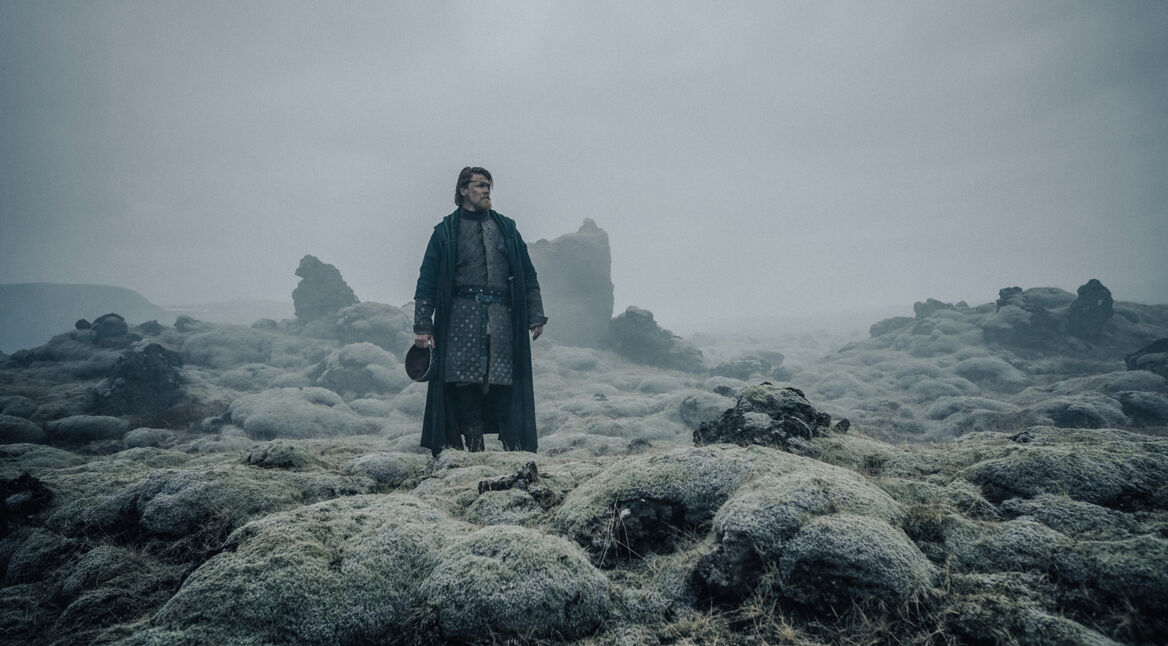 In dieser Szene aus "Hagen Im Tal der Nibelungen" steht Hagen, gespielt von Gísli Örn Garðarsson, allein in einer kargen, nebligen Landschaft voller moosbedeckter Felsen. Er trägt einen langen, mittelalterlichen Mantel und hält seinen Helm in der Hand. Die Umgebung wirkt düster und mystisch, verstärkt durch den grauen Himmel und die raue, unbewohnte Landschaft. Hagens ernste Miene und Haltung verleihen der Szene eine nachdenkliche und dramatische Atmosphäre.