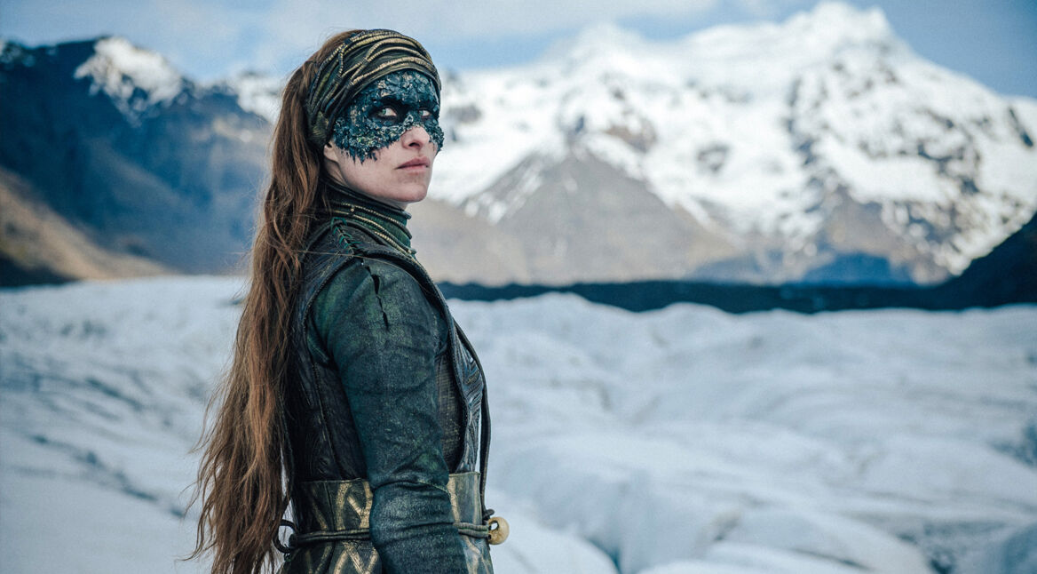 Auf dem Bild aus "Hagen im Tal der Nibelungen" ist Brunhild, die Königin von Island, gespielt von Rosalinde Mynster, zu sehen. Sie trägt ein grünes, detailreiches Kostüm und eine verzierte Maske. Im Hintergrund erstreckt sich eine schneebedeckte Berglandschaft, die die kühle und mystische Atmosphäre der Szene betont.