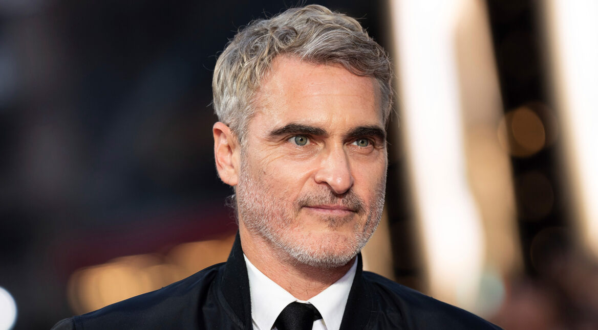 Auf dem Bild zu "Joaquin Phoenix Fakten" ist der Schauspieler Joaquin Phoenix in einer Nahaufnahme zu sehen. Er trägt ein schwarzes Jackett mit weißem Hemd und schwarzer Krawatte. Sein kurzes, graumeliertes Haar und der leicht graue Bart verleihen ihm ein reifes und markantes Aussehen. Der Hintergrund ist unscharf, mit warmen Lichtern, was die Konzentration auf sein Gesicht lenkt. Sein Blick ist ruhig und leicht fokussiert.
