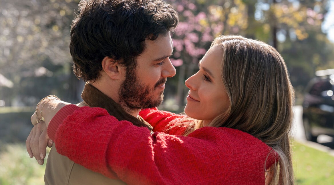 Auf dem Bild aus der Serie „Nobody Wants This“ sind die Charaktere Joanne und Noah in einer liebevollen Umarmung zu sehen. Sie schauen sich mit zärtlichen Blicken an, während Joanne ihre Arme um Noah legt. Beide lächeln sich sanft an, was eine tiefe Verbundenheit zwischen ihnen vermittelt. Joanne trägt einen roten Pullover, und im Hintergrund blühen Bäume in einer friedlichen, sonnigen Parklandschaft, was die warme und romantische Atmosphäre der Szene unterstreicht.