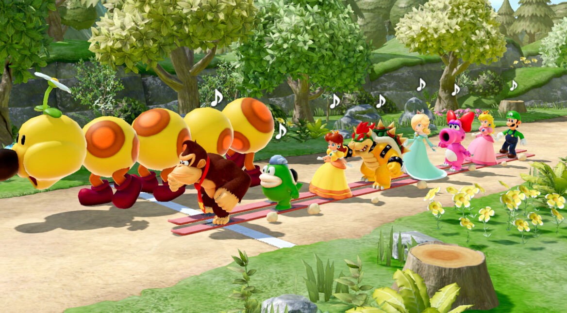 Auf diesem Bild aus dem Super Mario Party-Jamboree-Test sieht man eine bunte Reihe von Nintendo-Charakteren, die auf einem Pfad hintereinander marschieren. Angeführt wird die Gruppe von Donkey Kong, gefolgt von Figuren wie Yoshi, Daisy, Bowser, Rosalina, Birdo, Peach und Luigi. Vor Donkey Kong läuft ein großer, gelber Wiggler mit fröhlichem Gesichtsausdruck. Die Umgebung ist eine grüne, lebendige Waldlandschaft mit Blumen und Bäumen, und Musiknoten über den Köpfen der Charaktere zeigen, dass sie im Rhythmus marschieren.
