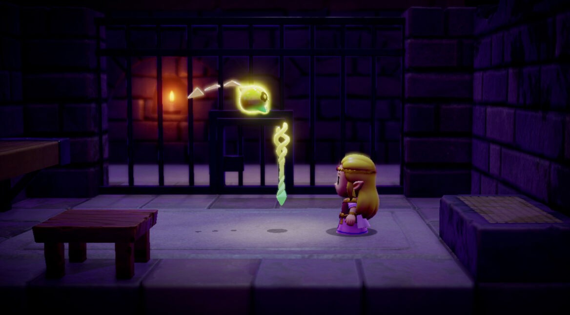 Auf dem Bild zu "Zelda: Echoes of Wisdom Tipps" ist Prinzessin Zelda in einer düsteren, steinernen Umgebung zu sehen. Vor ihr schwebt der leuchtende Tri-Sta, ein wichtiger Gegenstand für den Spielfortschritt. Im Hintergrund befinden sich Gitterstäbe und eine brennende Fackel, die die Atmosphäre verstärken. Zelda trägt ein lilafarbenes Kleid und eine goldene Krone, während sie den Tri-Sta konzentriert betrachtet.