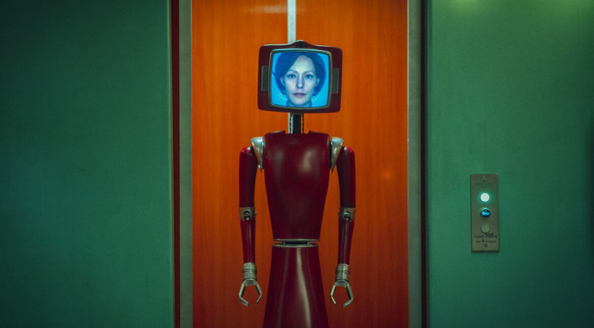 Auf dem Bild aus "Cassandra" ist Lavinia Wilson als Cassandra in Form eines humanoiden Roboters dargestellt. Der Roboter hat einen roten, metallischen Körper mit Greifarmen und einen Bildschirm als Kopf, auf dem das Gesicht von Cassandra zu sehen ist. Der Hintergrund zeigt einen orangefarbenen Aufzug und grüne Wände, was eine futuristische, aber auch retroartige Atmosphäre schafft.