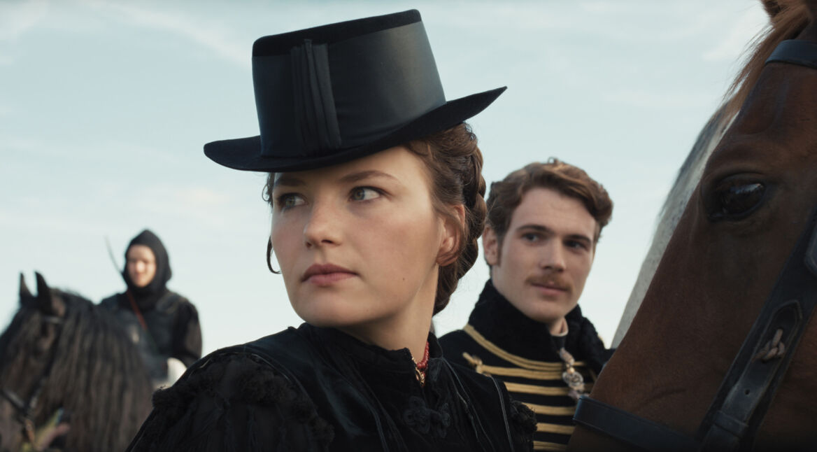 Das Bild zeigt Devrim Lingnau in ihrer Rolle als Kaiserin Elisabeth in der Serie "Die Kaiserin" an einem der Drehorte. Sie trägt einen eleganten schwarzen Reithut und ein passendes Kleid, während sie auf einem Pferd sitzt und ernsthaft in die Ferne blickt. Im Hintergrund sind ein weiterer Reiter und ein Mann in einer Uniform mit goldenen Verzierungen zu sehen, die die historische Szenerie unterstreichen.