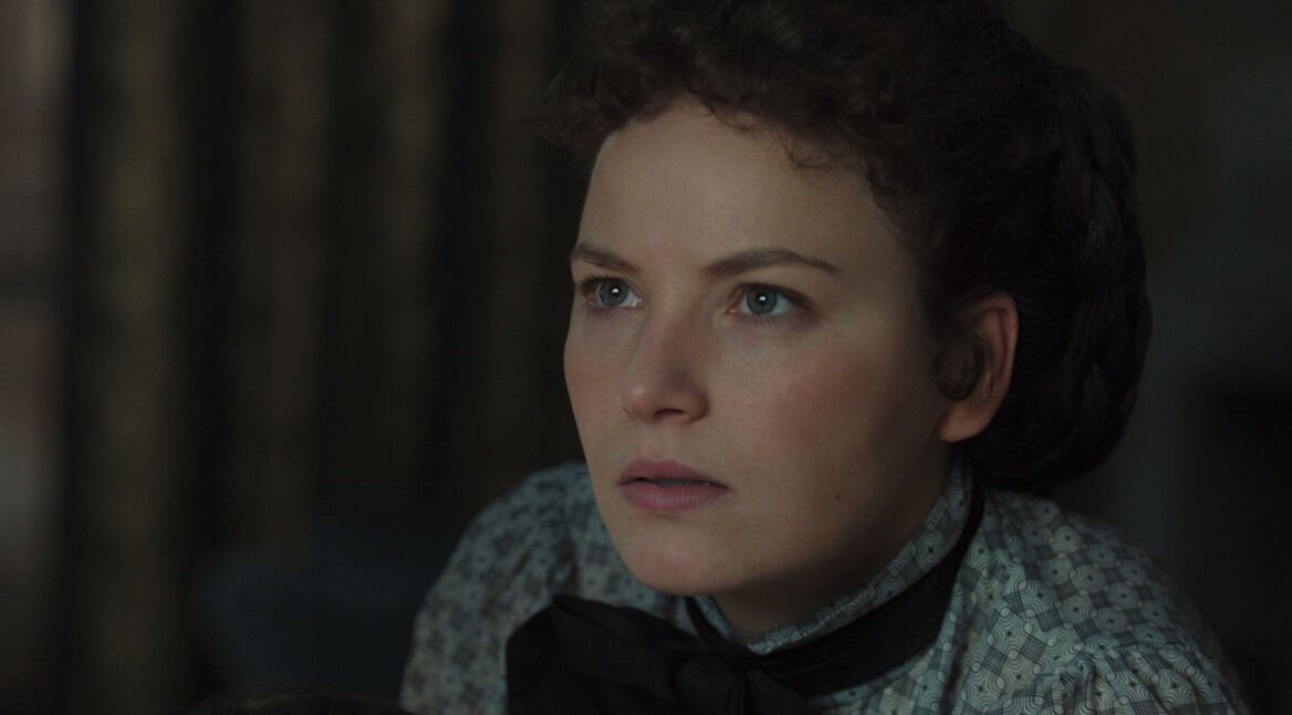 Auf dem Bild zur Serie "Die Kaiserin Staffel 2 Ende erklärt" ist Devrim Lingnau als Kaiserin Elisabeth, bekannt als Sisi, zu sehen. Ihr Gesichtsausdruck ist ernst und konzentriert, was auf eine emotionale oder bedeutsame Szene hinweist. Sie trägt eine dezente, gemusterte Bluse mit einer schwarzen Schleife, die ihren klassischen Stil unterstreicht. Der Hintergrund ist dunkel und betont die Intensität des Moments. Die Szene vermittelt Nachdenklichkeit und eine mögliche innere Auseinandersetzung der Figur.