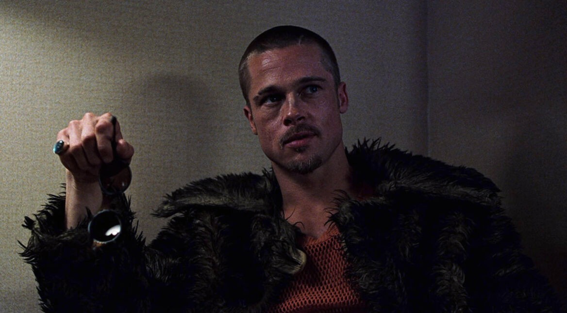 Auf dem Bild zu Fight Club Zitate ist Brad Pitt als Tyler Durden zu sehen. Er trägt eine dunkle, flauschige Jacke und ein rotes Netzshirt, sein Blick wirkt intensiv und herausfordernd. In seiner rechten Hand hält er eine Sonnenbrille, während er leicht seitlich sitzt und nachdenklich dreinschaut.