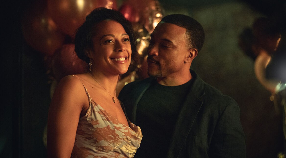 Auf dem Bild zur Serie "Ich vermisse Dich" sind Rosalind Elezar und Ashley Walters zu sehen. Sie stehen eng beieinander, Rosalind lächelt strahlend, während Ashley sie zärtlich anschaut. Im Hintergrund sind Ballons und ein warmes, schummriges Licht zu erkennen, das eine intime und herzliche Atmosphäre schafft.