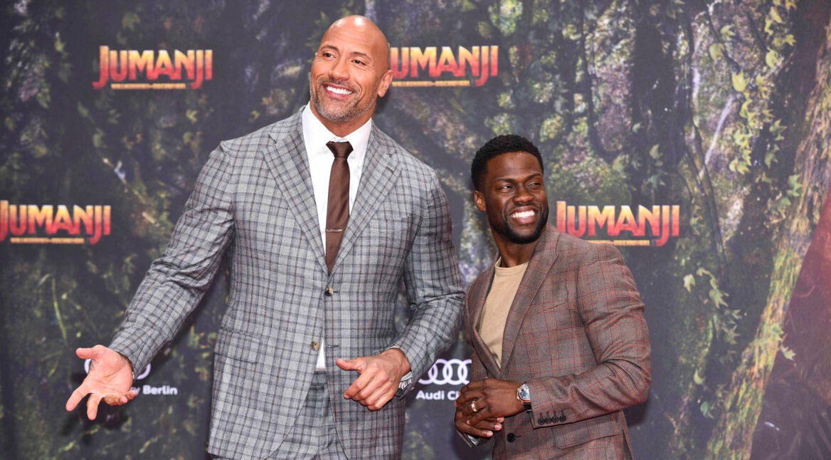 Auf dem Bild zu Jumanji 3 sind Dwayne Johnson (links) und Kevin Hart (rechts) gemeinsam auf dem roten Teppich des Vorgängerfilms zu sehen. Johnson trägt einen grauen karierten Anzug und lächelt breit, während Hart ebenfalls im karierten Anzug lächelt und zu ihm hochblickt. Der Hintergrund zeigt das Jumanji-Logo vor einer Dschungelkulisse.