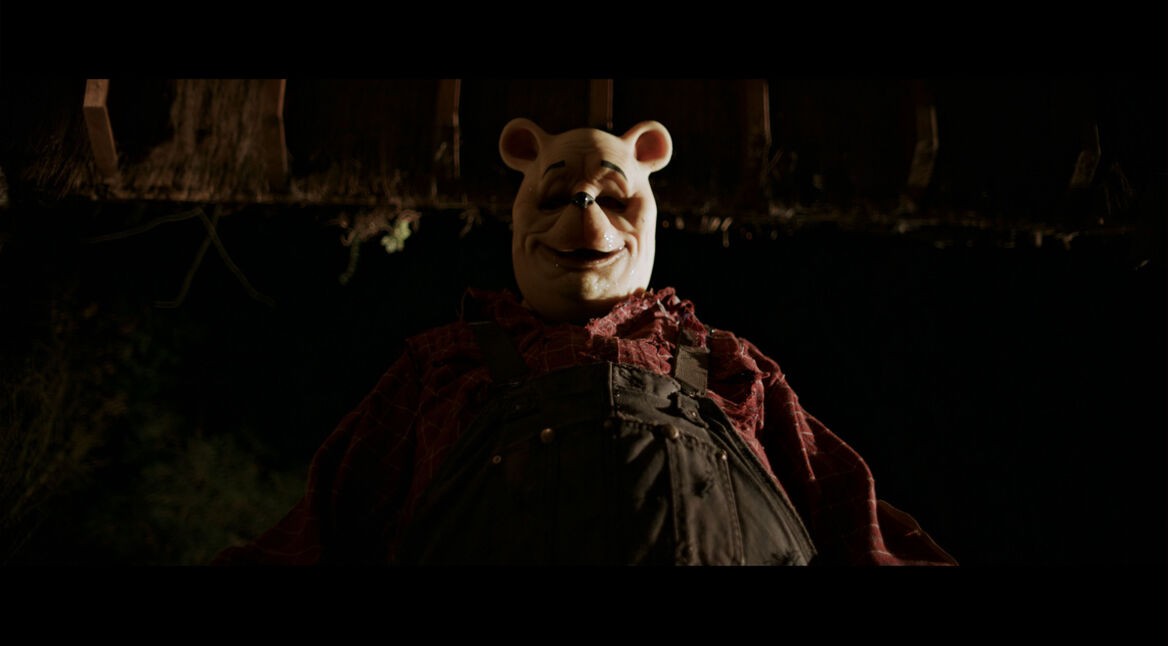 Dieses Bild aus dem Film Winnie the Pooh: Blood and Honey zeigt eine düstere, unheimliche Version von Winnie Puuh, die das klassische Kinderbuch-Image ins Horror-Genre überführt. Die Figur trägt eine beunruhigende Maske mit markanten, kindlichen Zügen, kombiniert mit abgenutzter Latzhose und kariertem Hemd. Die Szene ist in dunklen, bedrohlichen Farben gehalten, was die gruselige Atmosphäre unterstreicht. Der Film ist der erste in einer Reihe von Kinderbuch-Horrorfilmen im Poohniverse.