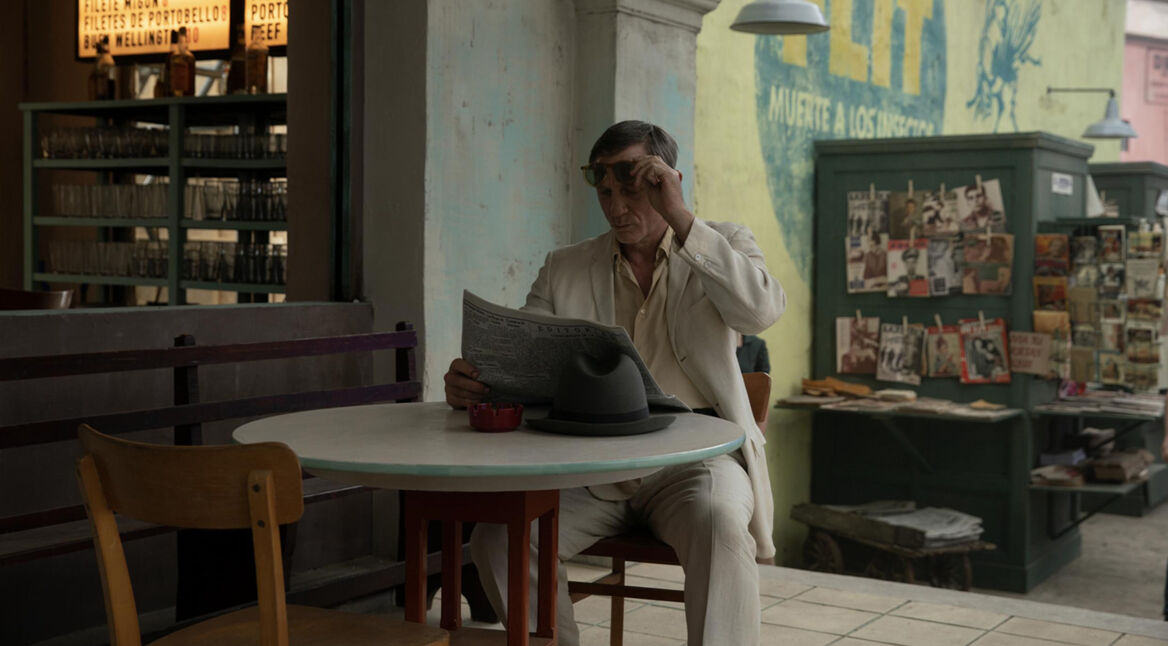 In dieser Szene aus dem Film Queer sitzt William Lee, gespielt von Daniel Craig, in einem Café. Er trägt einen hellen Anzug, liest eine Zeitung und hebt seine Sonnenbrille leicht an. Auf dem Tisch liegt sein Hut, daneben steht ein Aschenbecher. Im Hintergrund sind ein Regal mit Gläsern und ein Zeitschriftenstand zu sehen, was eine nostalgische Atmosphäre erzeugt.