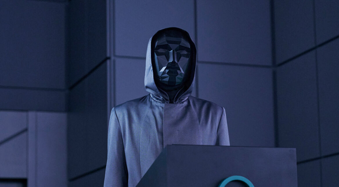 Das Bild im Artikel zum Squid Game US-Remake zeigt eine Szene aus der zweiten Staffel der Originalserie, in der der Frontman, eine mysteriöse Figur in einem kantigen, schwarzen, geometrischen Maske und einem grauen Kapuzenmantel, hinter einem Pult steht, umgeben von einer düsteren, blau beleuchteten Umgebung.