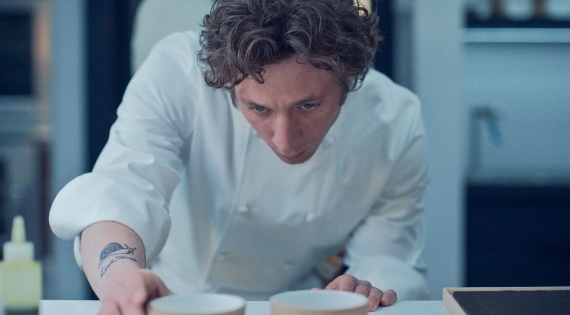 Auf dem Bild zur Serie The Bear Staffel 4 ist Carmen „Carmy“ Berzatto, gespielt von Jeremy Allen White, zu sehen. Er trägt eine weiße Kochjacke und ist konzentriert, während er zwei Schalen auf einem Tisch präzise arrangiert. Sein Blick ist fokussiert, und ein Schnecken-Tattoo ist auf seinem Unterarm sichtbar. Die Szene vermittelt eine intensive Arbeitsatmosphäre in einer professionellen Küche.