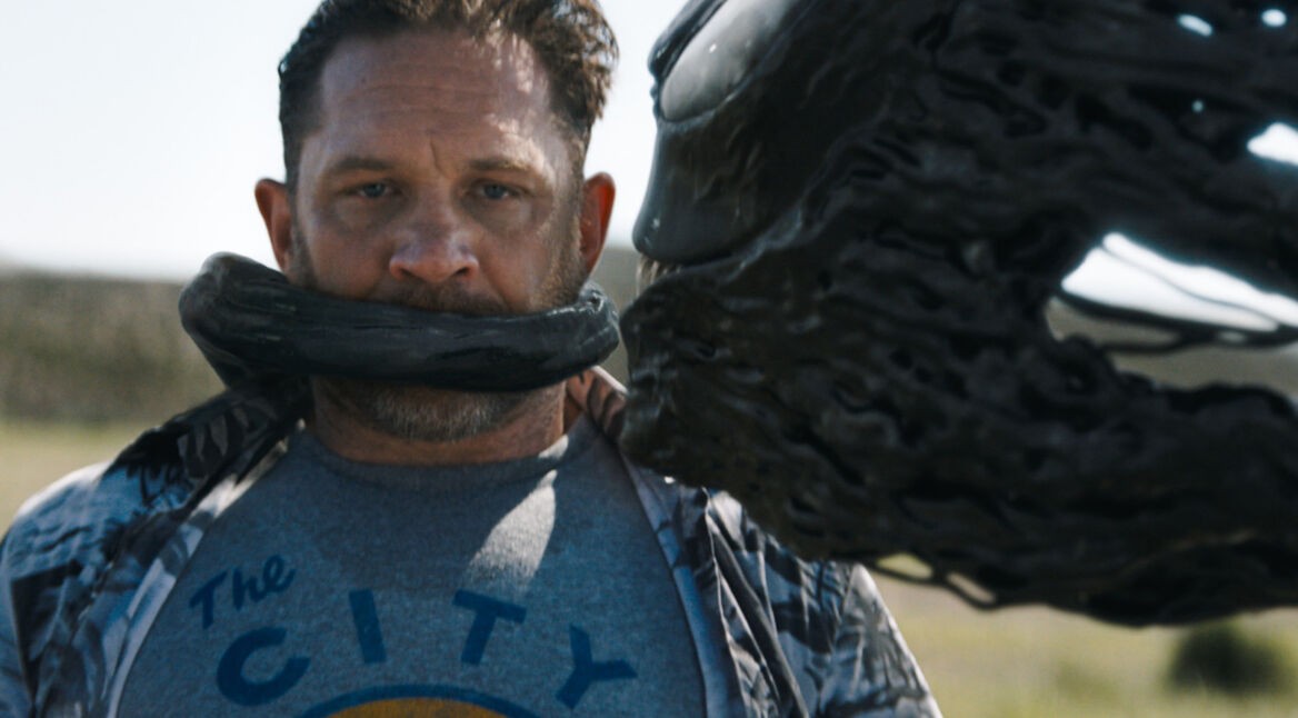 In dieser Szene aus Venom 3 ist Tom Hardy als Eddie Brock zu sehen, wie ihn der schwarze, flüssige Symbiont Venom teilweise umhüllt und seinen Mund bedeckt. Er trägt ein graues T-Shirt mit der Aufschrift „The City“ und blickt ernst nach vorne. Die Umgebung im Hintergrund ist verschwommen, was den Fokus auf die bedrohliche Verbindung zwischen Brock und Venom legt.