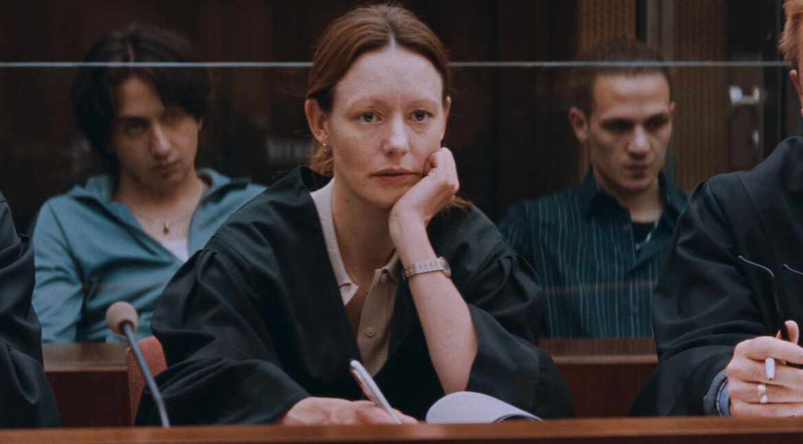 Auf dem Bild in dem Artikel über die Zeit Verbrechen-Serie, ist eine Frau mit roten Haaren zu sehen, die wie eine Anwältin gekleidet ist. Im Hintergrund sind weitere Menschen zu sehen. Der Raum in dem sie sitzen scheint ein Gerichtssaal zu sein.