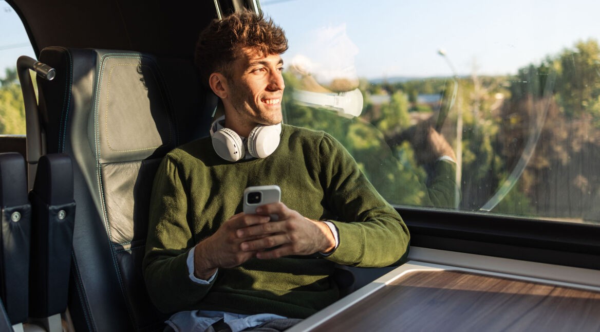Ein Mann sitzt mit einem Smartphone und Kopfhörern im Zug und schaut aus dem Fenster