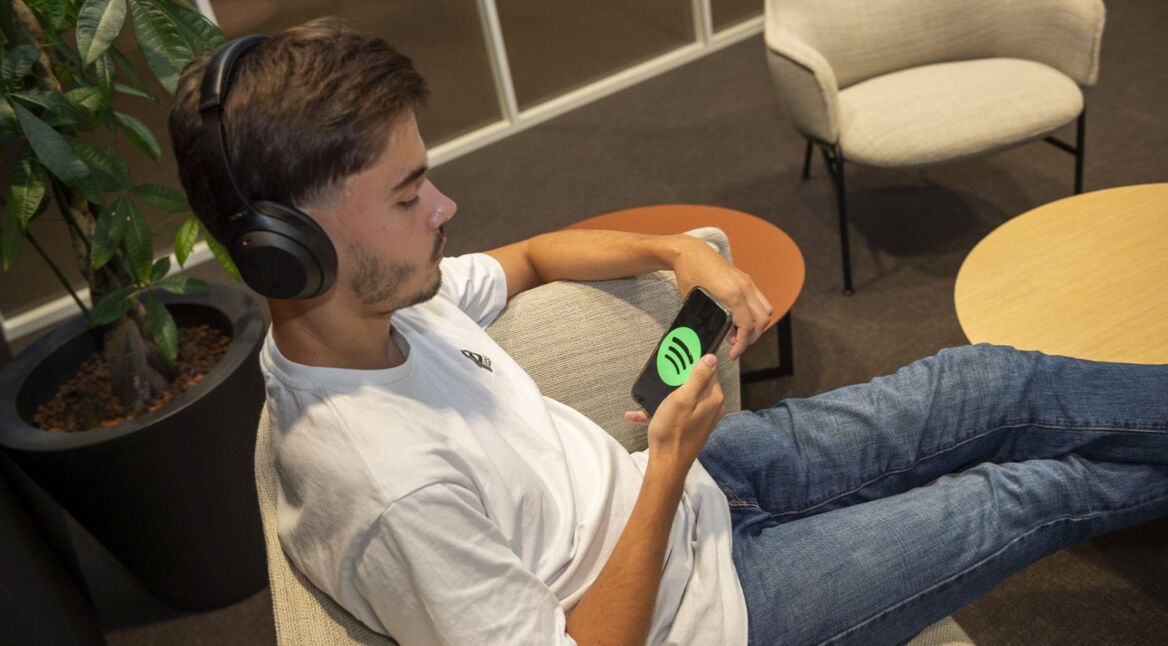 Junger Mann mit Kopfhörern sitzt auf Couch und hört am Smartphone Musik via Spotify