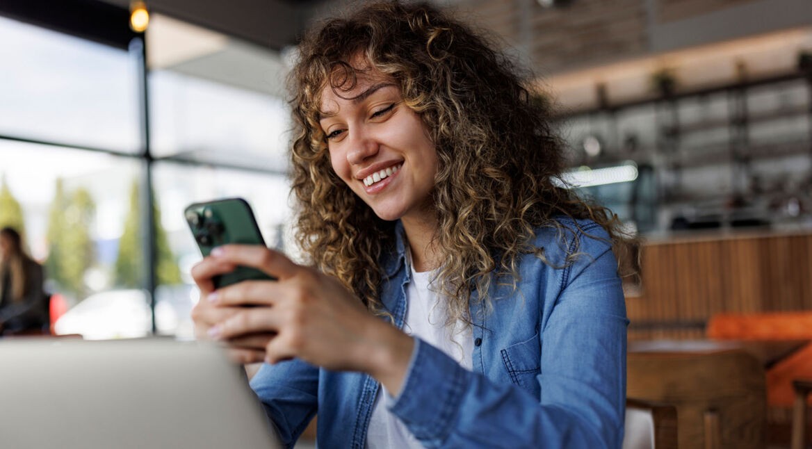 Eine junge Frau sitzt mit einem Handy in einem Café und lächelt.