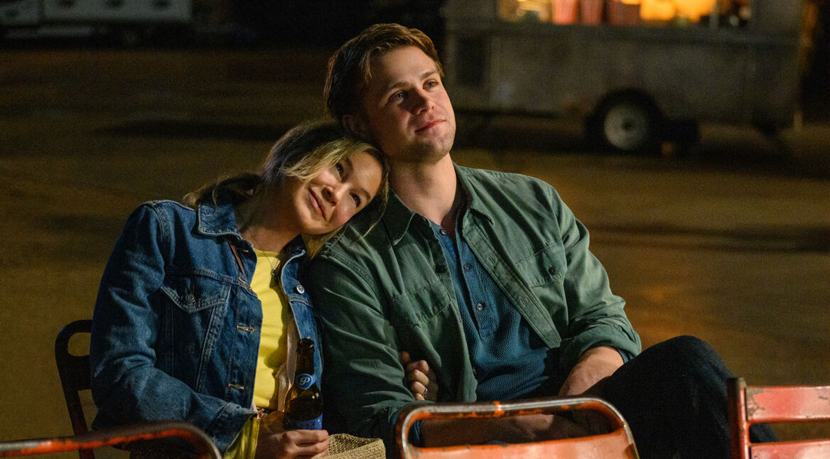 Auf dem Bild aus Bridget Jones 4 sind Bridget Jones, gespielt von Renée Zellweger, und Roxter, dargestellt von Leo Woodall, zu sehen. Bridget lehnt sich entspannt an Roxters Schulter, beide wirken ruhig und zufrieden. Sie sitzen in einer nächtlichen Outdoor-Szene auf einfachen Stühlen, während Bridget eine Bierflasche in der Hand hält. Im Hintergrund ist eine unscharfe Umgebung mit einem Anhänger und warmem Licht zu erkennen.