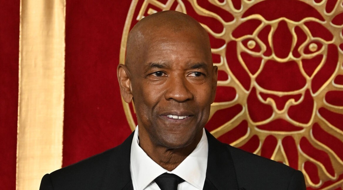 Auf dem Bild zu Denzel Washingtons besten Filmmomenten ist Denzel Washington in einem eleganten schwarzen Anzug mit weißem Hemd und schwarzer Krawatte zu sehen. Er lächelt dezent, während er vor einem roten Hintergrund mit goldenen Verzierungen, die ein Löwenmotiv zeigen, steht. Die Szene strahlt eine feierliche und stilvolle Atmosphäre aus.