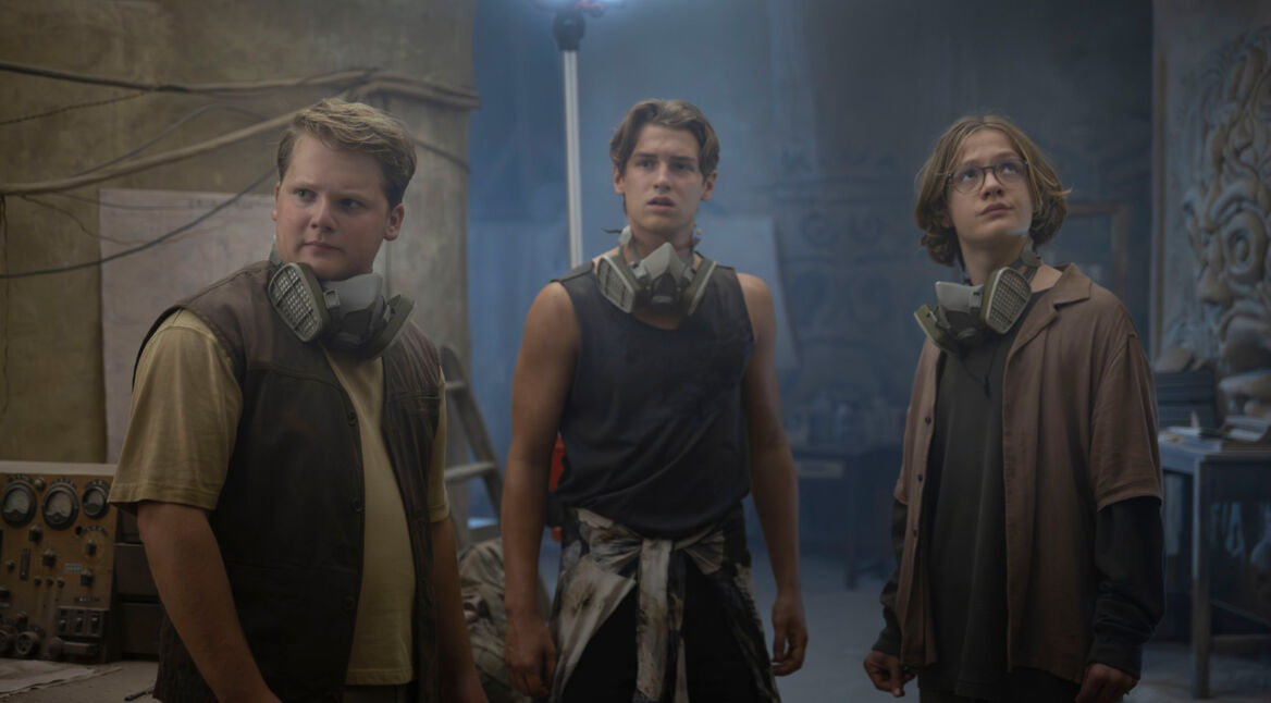 In „Die drei ??? – Toteninsel“ stehen Justus Jonas (Julius Weckauf), Peter Shaw (Nevio Wendt) und Bob Andrews (Levi Brandl) in einem staubigen, mysteriös beleuchteten Raum. Sie tragen alle Atemschutzmasken um den Hals und wirken alarmiert, während sie sich aufmerksam umsehen.