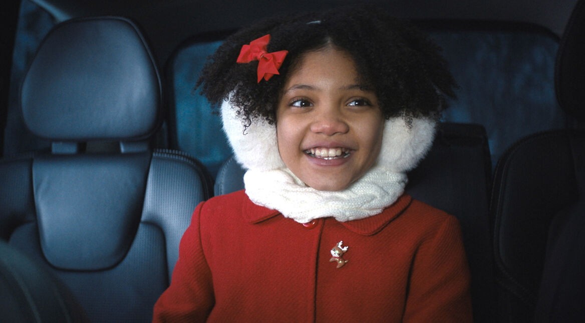 Auf dem Bild im GigaTV-Highlights Dezember 2024-Artikel siehst Du Leah Brady als Trudy in Violent Night. Sie sitzt lächelnd in einem Auto, trägt eine rote Jacke mit einer Rentier-Brosche, einen weißen Schal und Ohrenschützer.