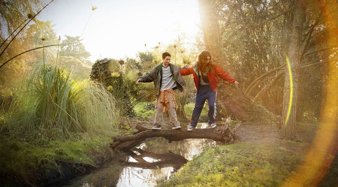Auf dem Visual zum Weihnachtsspot von Vodafone 2024 balancieren zwei Jugendliche fröhlich auf einem umgestürzten Baumstamm über einen kleinen Bach in einer idyllischen Naturkulisse. Sie tragen lässige, bunte Kleidung, während goldenes Sonnenlicht durch die Bäume scheint und eine warme, festliche Stimmung erzeugt.