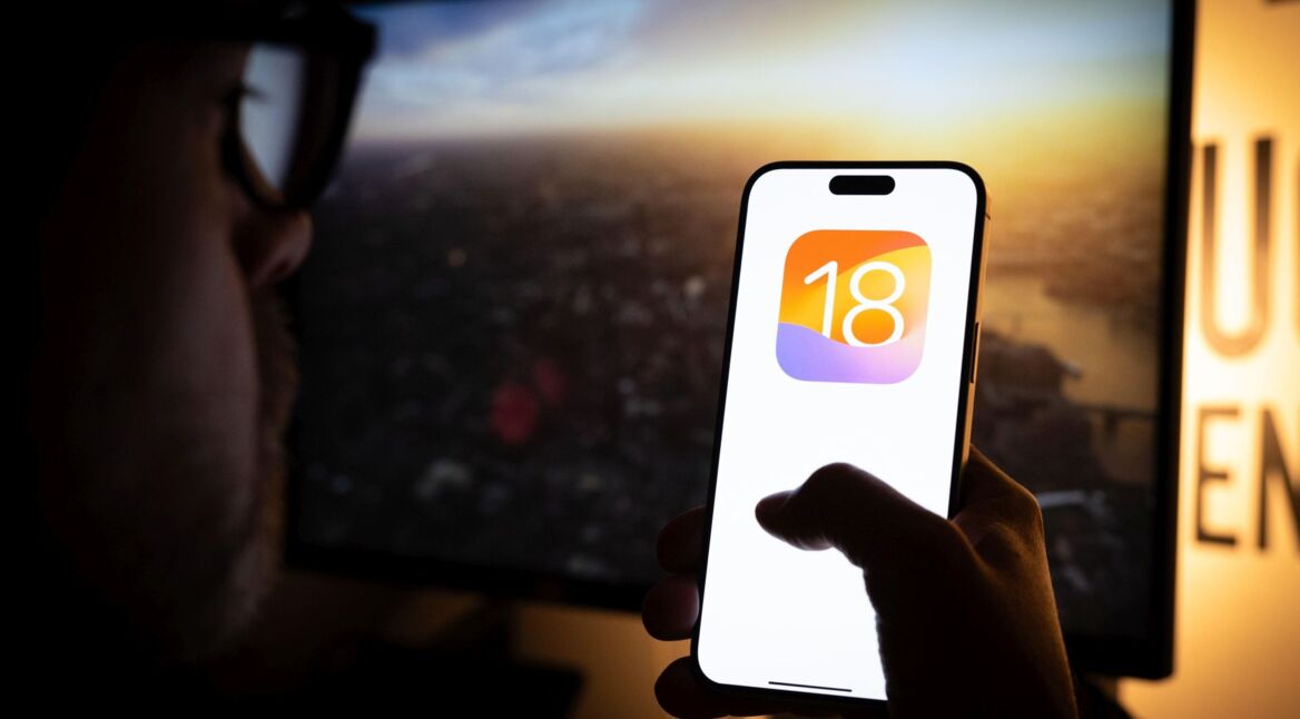 Das Logo von iOS 18 ist auf einem iPhone zu sehen, das ein junger Mann mit Brille hochhält.