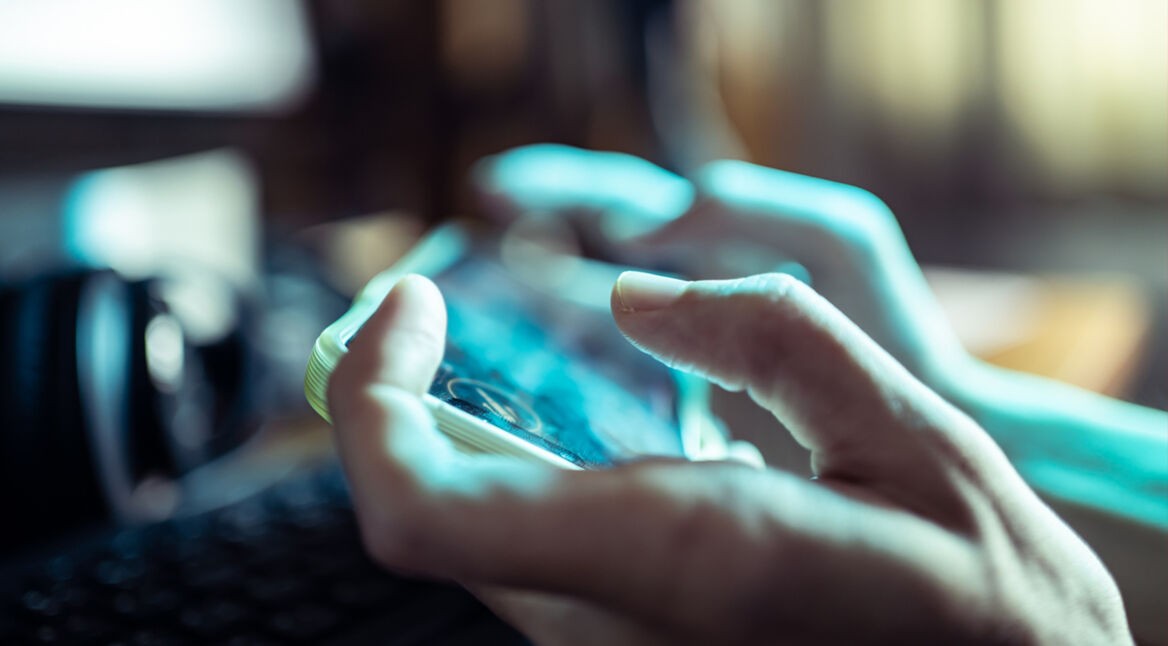 Das Bild in dem Artikel über die besten Koop-Games fürs Handy, zeigt eine Nahaufnahme von Händen, die ein Smartphone im Querformat halten. Der Fokus liegt auf den Fingern und dem Bildschirm, der unscharf, aber lebendig wirkt. Die Atmosphäre ist konzentriert, das Spielgeschehen spiegelt sich leicht auf dem Display wider.