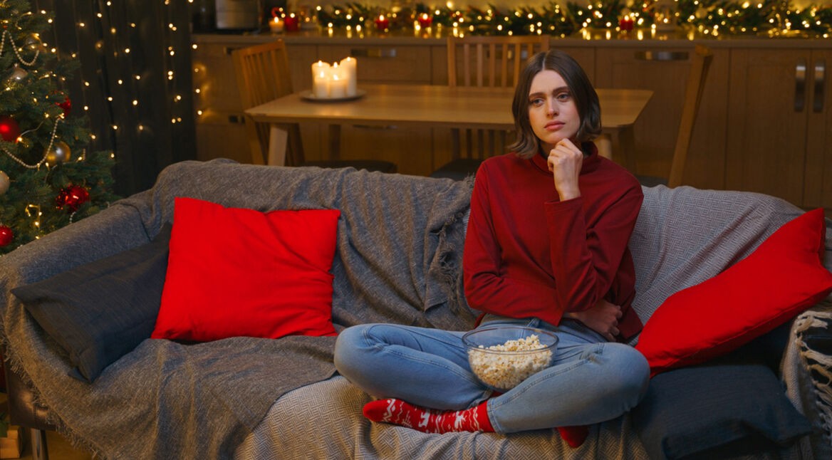 Eine junge Frau in einem roten Pullover sitzt auf einem grauen Sofa, umgeben von roten Kissen, und hält eine Schüssel Popcorn. Sie wirkt nachdenklich und leicht gelangweilt. Im Hintergrund ist ein festlich geschmückter Weihnachtsbaum mit Lichtern und roten Kugeln zu sehen, zusammen mit Kerzen und Lichterketten. Perfekt passend zum Thema "Schlechter Weihnachtsfilm".