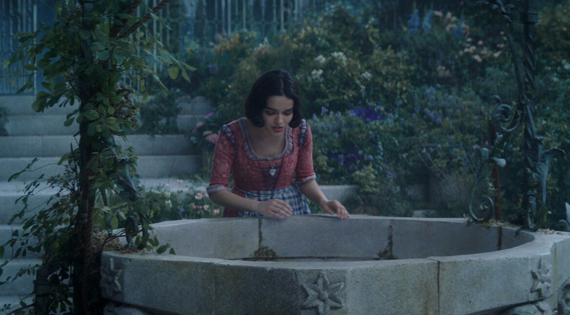 In einer Szene aus der Schneewittchen Realverfilmung ist Schneewittchen an einem steinernen Brunnen zu sehen. Sie trägt ein rotes Oberteil mit blauem Kleid und beugt sich über den kunstvoll verzierten Brunnenrand.